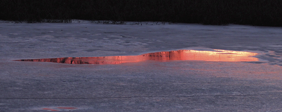 “On a small frozen pond the setting sun can produce dramatic reflections in surface melt water. The mirror-like surface of the water and its irregular shape are surrounded by the cold blue non-reflective surface of the ice. It is a fleeting event as the sunset sky fades into twilight.” – Carl Kurtz