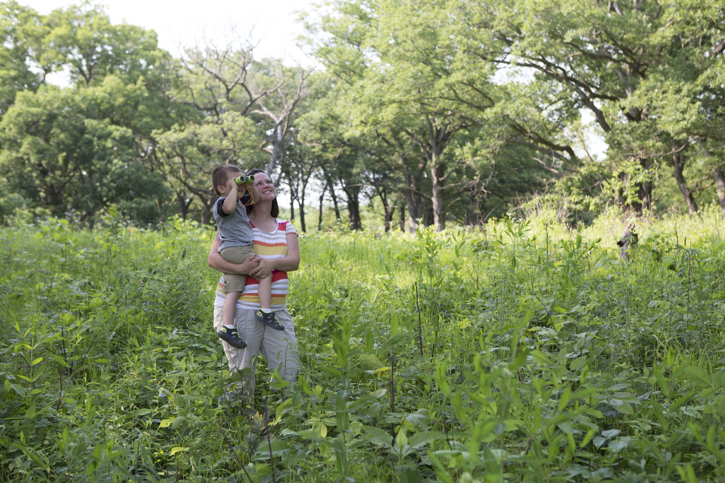 A mother holds her child outside as he uses binoculars.