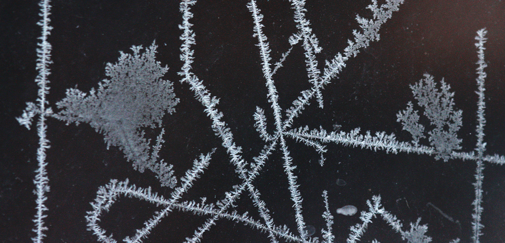 Everyone in cold climates has likely seen frost patterns on a window after a very cold night.  It can take many different forms or shapes from lines to flowing flower-like designs.  Formation occurs due to the difference in very cold air outside and moist humid air inside.  Imperfections in the glass or on its surface such as dust, dirt, or scratches determine the form of the crystals.  Higher humidity will also affect the thickness of the ice. 